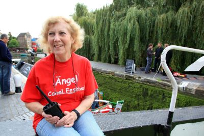 A middle aged lady in red festival sweatshirt seated on a lock gate beam, smiling, holding radio