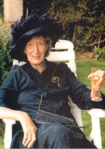 An older woman in a dark dress and hat sits in a chair