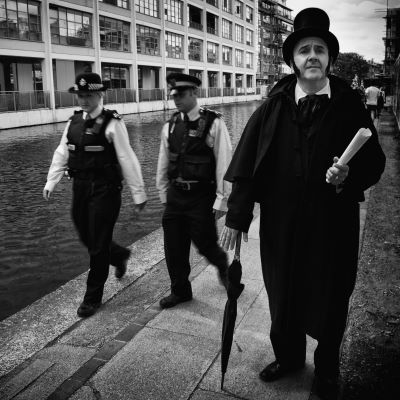 A man in a dark costume and top had walks towards the camera as two police offers follow