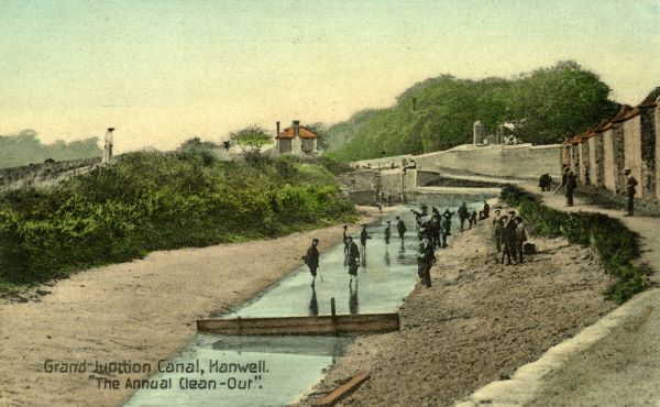 A canal is seen dry, with workers in it clearing things, large walll to the right